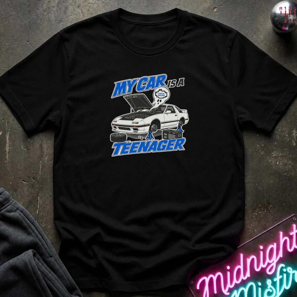 A black short-sleeve graphic t-shirt laid flat on a dark textured surface. The graphic features a white sports car with its hood open, blue text reading 'MY CAR IS A TEENAGER', and a small blue-and-white character above the car.