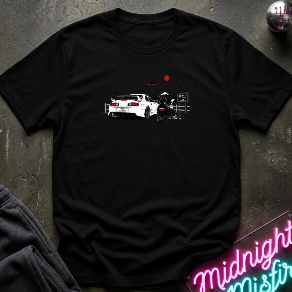 A black short-sleeve graphic T-shirt laid flat on a dark textured surface. The shirt features a printed illustration of a white sports car with a red sun and mountain graphic above it.