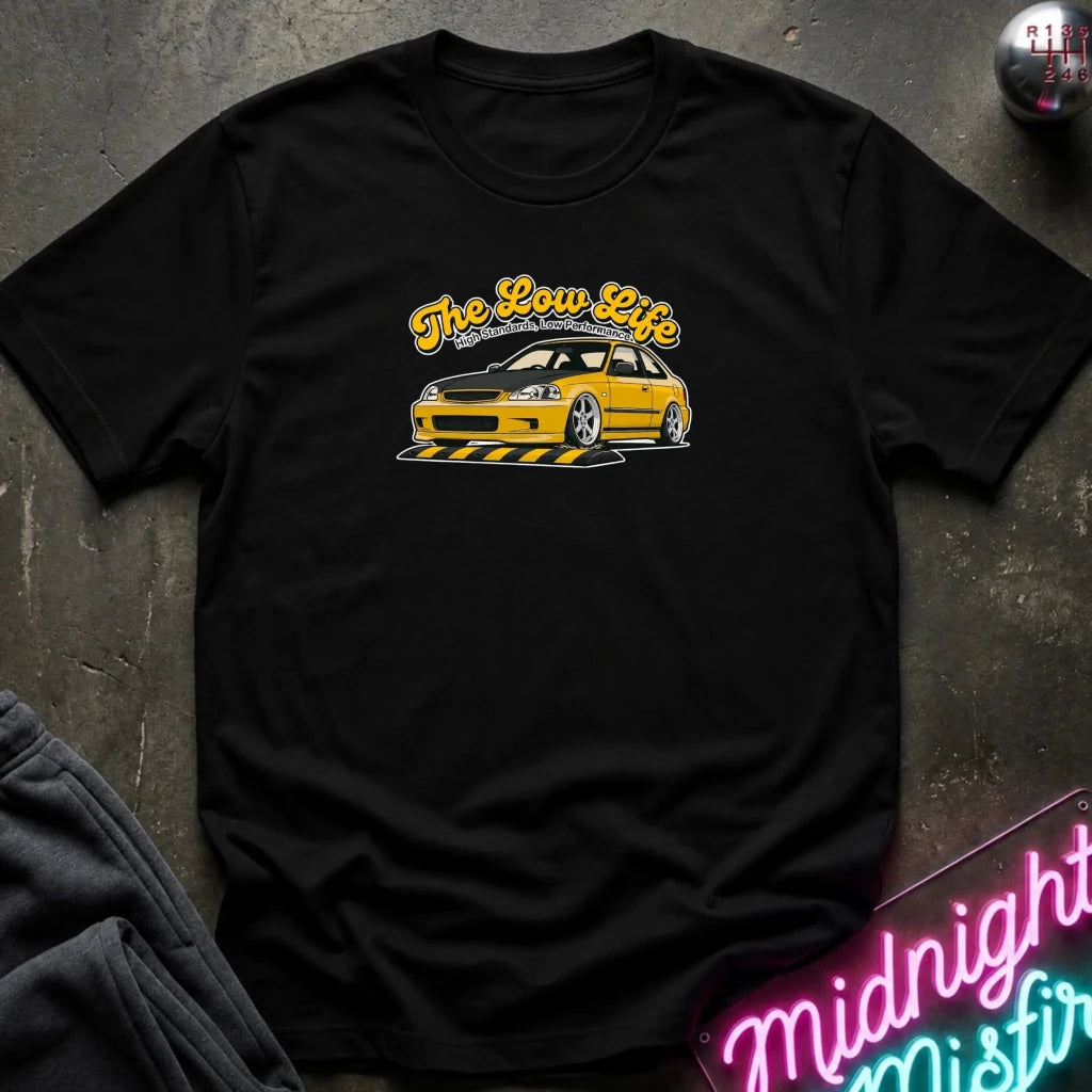 A black short-sleeve graphic t-shirt laid flat on a dark textured surface. The shirt features a yellow car and the text 'The Low Life' in yellow script with a white outline across the chest.