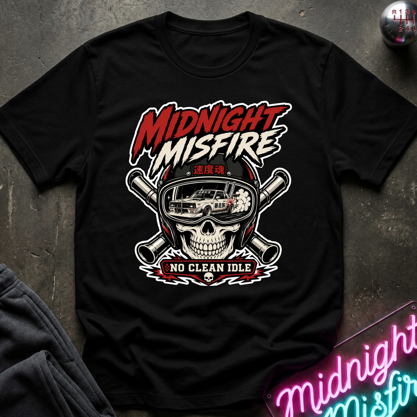 A black short-sleeve graphic T-shirt laid flat on a dark textured surface. The shirt features a large front print with the words “MIDNIGHT MISFIRE” in red and white, a skull wearing a racing helmet with a car inside, crossed pipes, and the phrase “NO CLEAN IDLE” below.