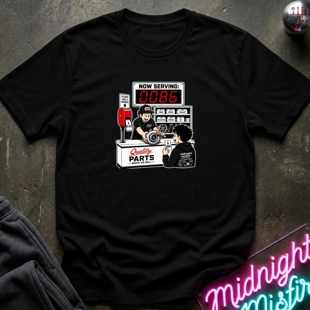 A black short-sleeve crewneck t-shirt featuring a retro-style graphic of a mechanic working at a parts counter with a digital display showing 'NOW SERVING: 0086'. 