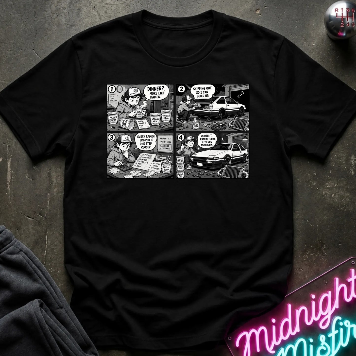 A black short-sleeve crewneck t-shirt featuring a black and white comic strip graphic on the chest, styled in a flat-lay on a dark textured surface with a neon sign and dark pants partially visible.