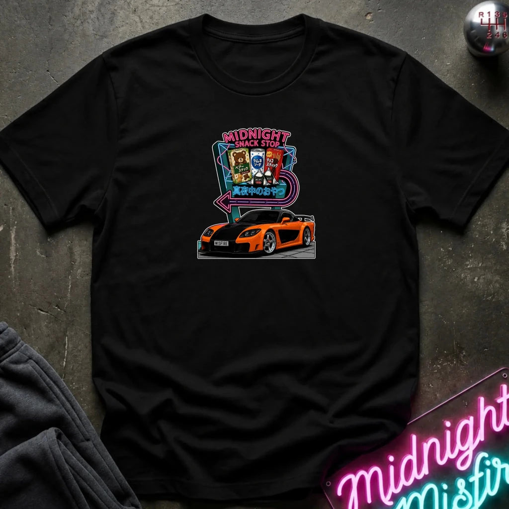 A black  short-sleeve t-shirt laid flat on a dark textured surface, featuring a colorful graphic print of an orange sports car in front of a neon sign that reads 'Midnight Snack Stop'.