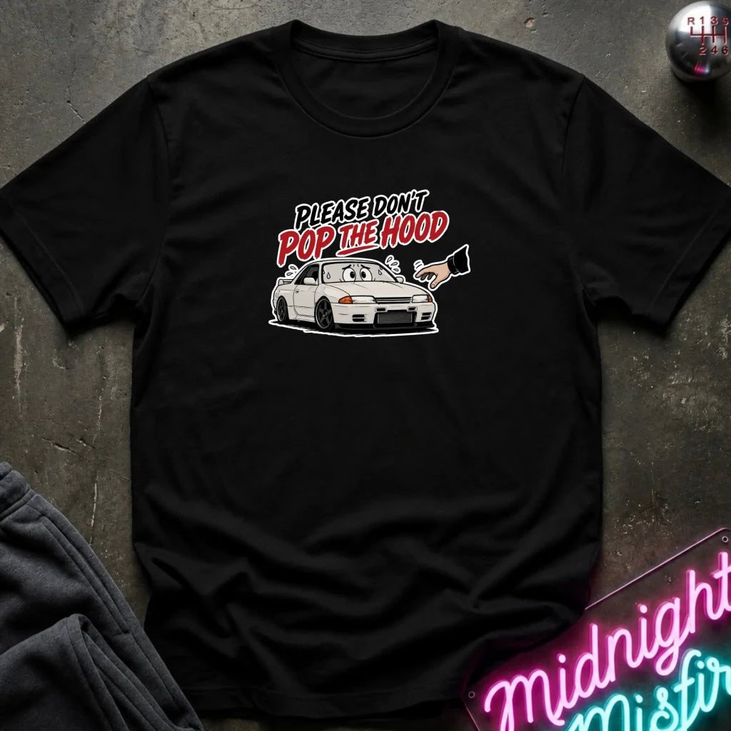 A black short-sleeve graphic t-shirt laid flat on a dark textured surface. The graphic features the text 'PLEASE DON'T POP THE HOOD' in white and red, above a white sports car with a hand holding a megaphone.
