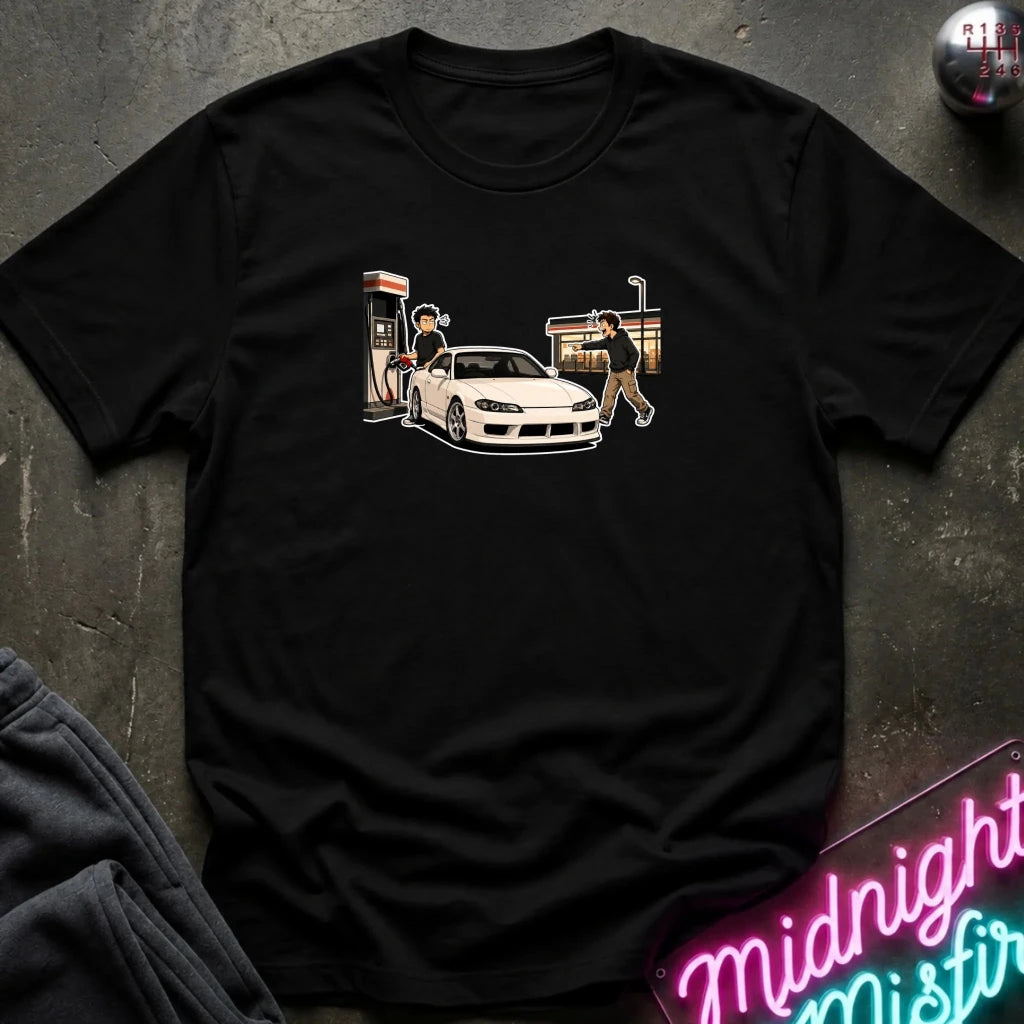 A black short-sleeve graphic t-shirt laid flat on a dark textured surface. The graphic shows a white sports car with a person standing next to it near a gas station pump and a convenience store.