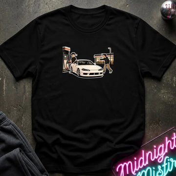 A black short-sleeve graphic t-shirt laid flat on a dark textured surface. The graphic shows a white sports car with a person standing next to it near a gas station pump and a convenience store.