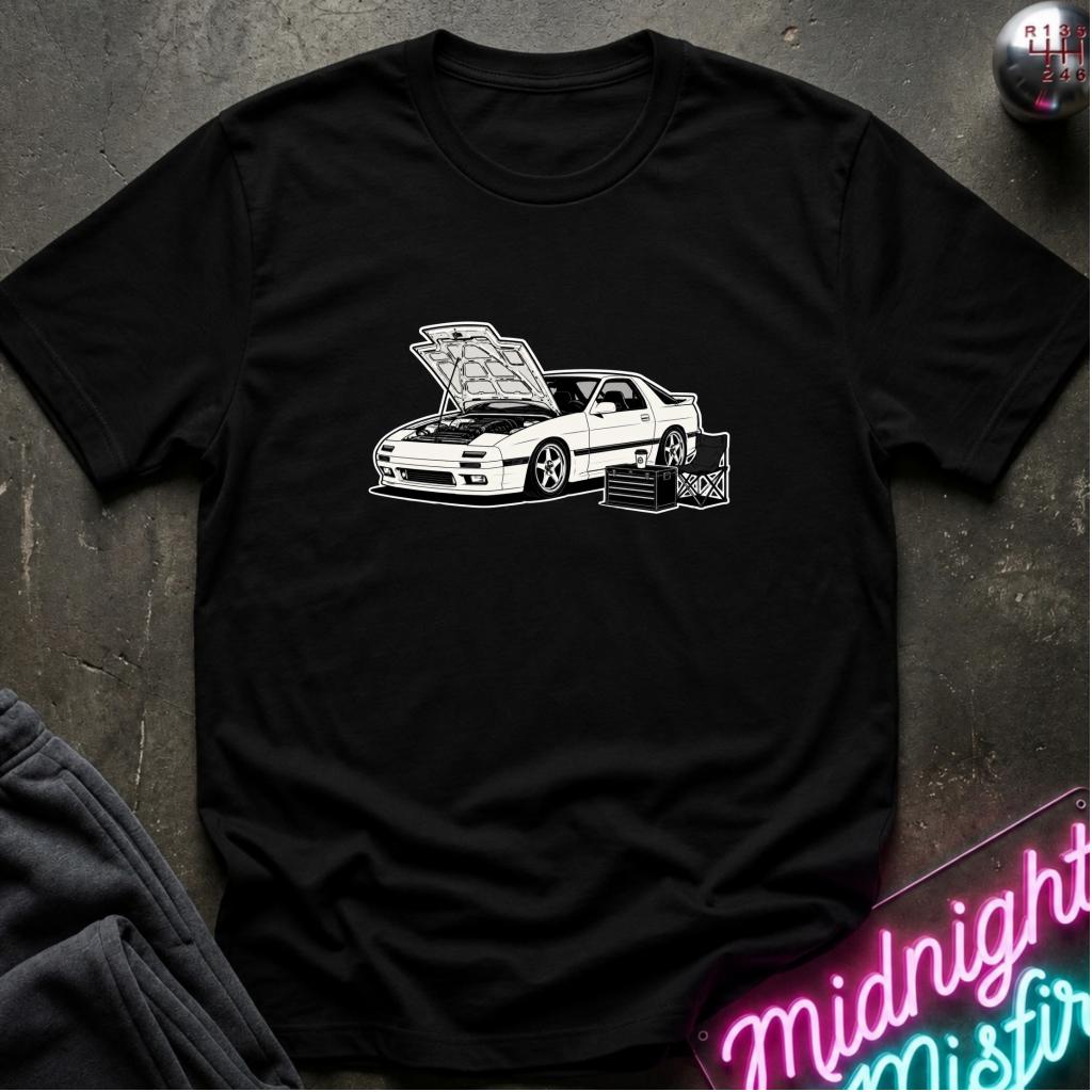 A black short-sleeve t-shirt laid flat on a dark textured surface, featuring a white line-art graphic of a sports car with its hood open and a toolbox beside it. A neon sign and folded dark fabric are partially visible around the edges as styling props.