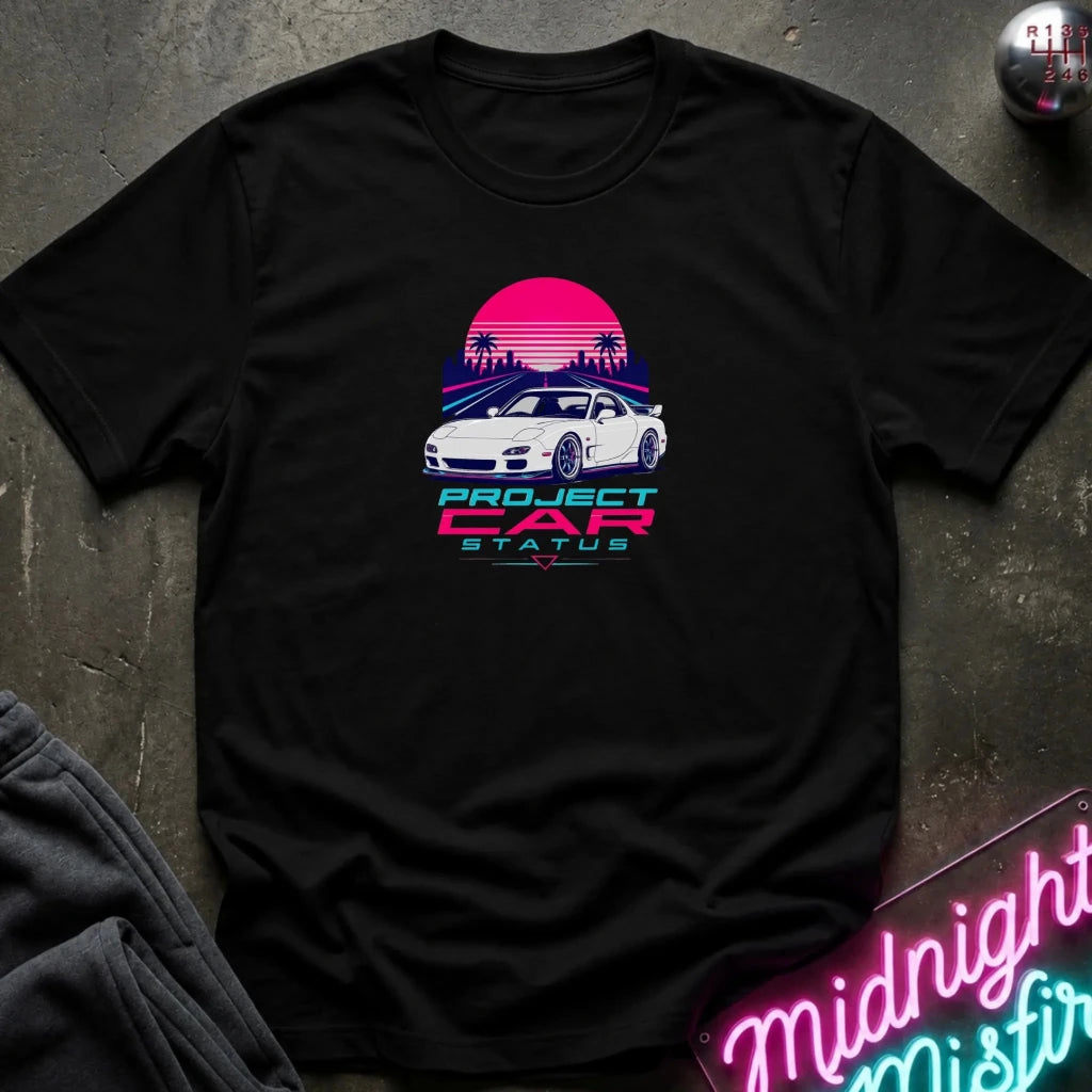 A black short-sleeve t-shirt laid flat on a dark textured surface, featuring a retro-style graphic of a white sports car in front of a pink and blue sunset with palm trees and the text 'PROJECT CAR STATUS'.