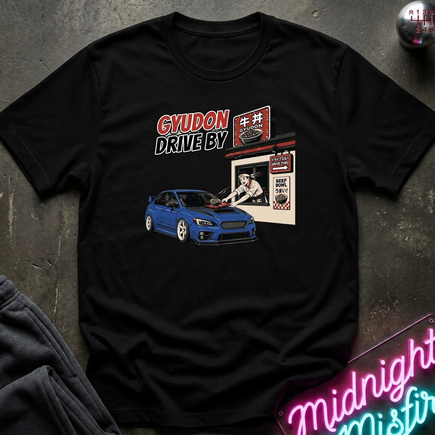 A black short-sleeve graphic t-shirt laid flat on a dark textured surface. The graphic features a blue sports car and a Japanese drive-in restaurant sign with the text 'GYUDON DRIVE BY'.