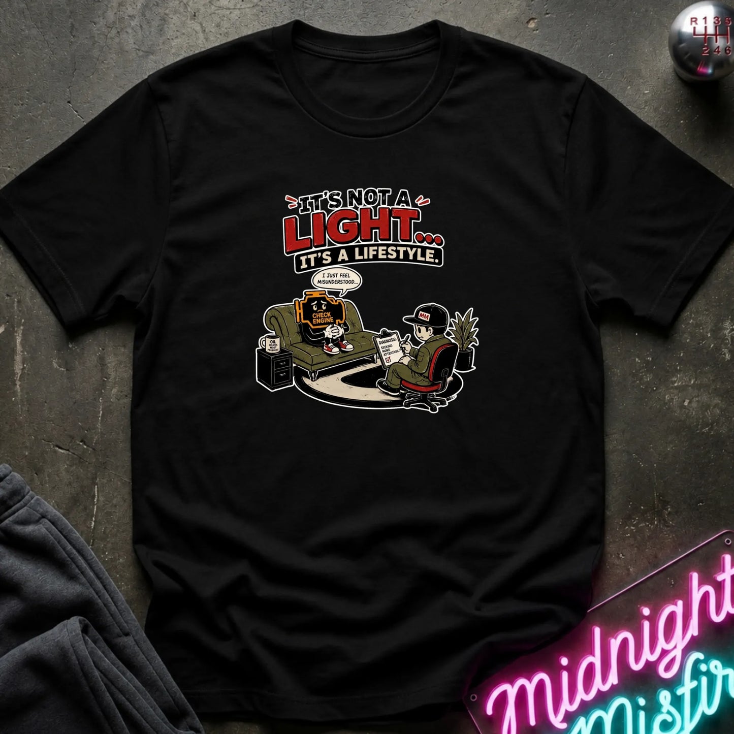 A black short-sleeve graphic t-shirt laid flat on a dark textured surface. The graphic features a cartoon character in a chair next to a person holding a pen, with the text 'IT'S NOT A LIGHT.
