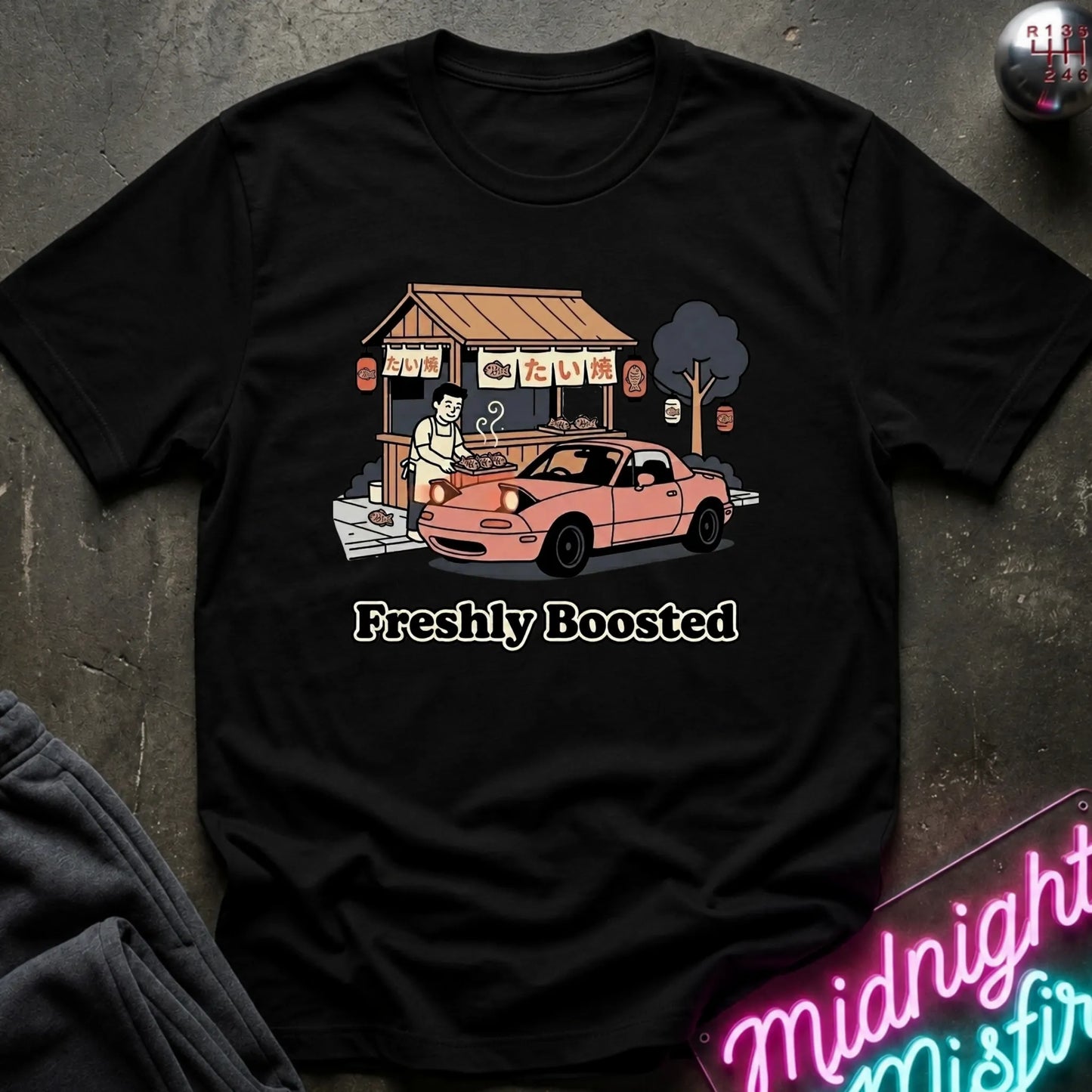 A black graphic t-shirt laid flat on a dark textured surface. The graphic features a pink convertible car parked in front of a street food stall  and the text 'Freshly Boosted' printed below.