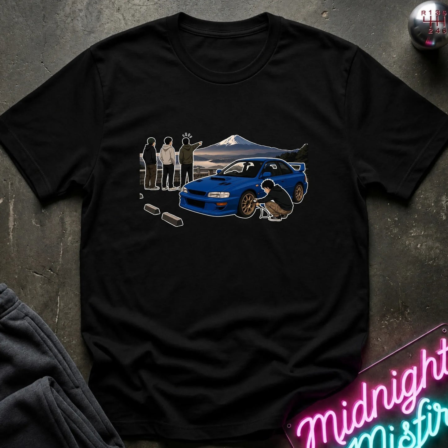 A black short-sleeve graphic t-shirt laid flat on a dark textured surface. The graphic shows a blue sports car, a mountain landscape, and several silhouetted figures.