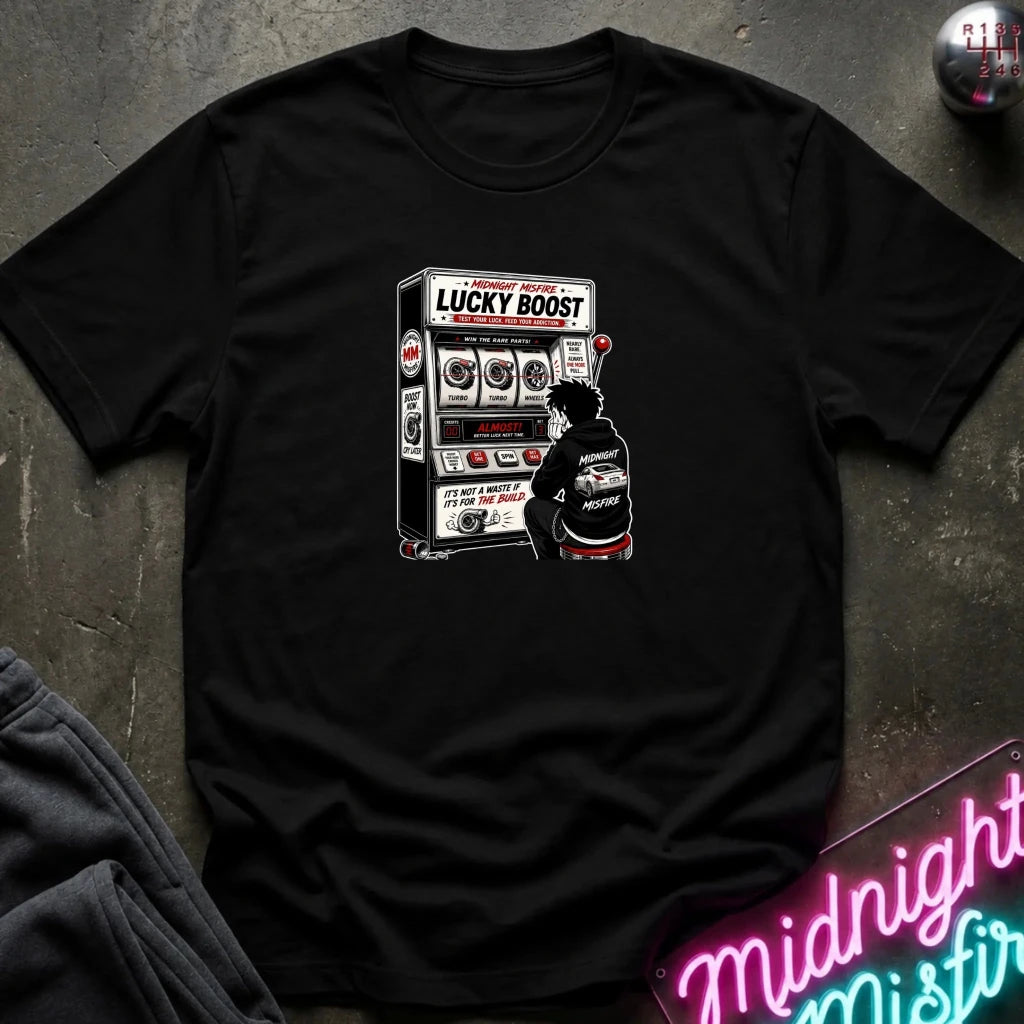 A black crewneck t-shirt laid flat on a dark textured surface, featuring a graphic print of a person sitting in front of a slot machine with the text 'LUCKY BOOST'.