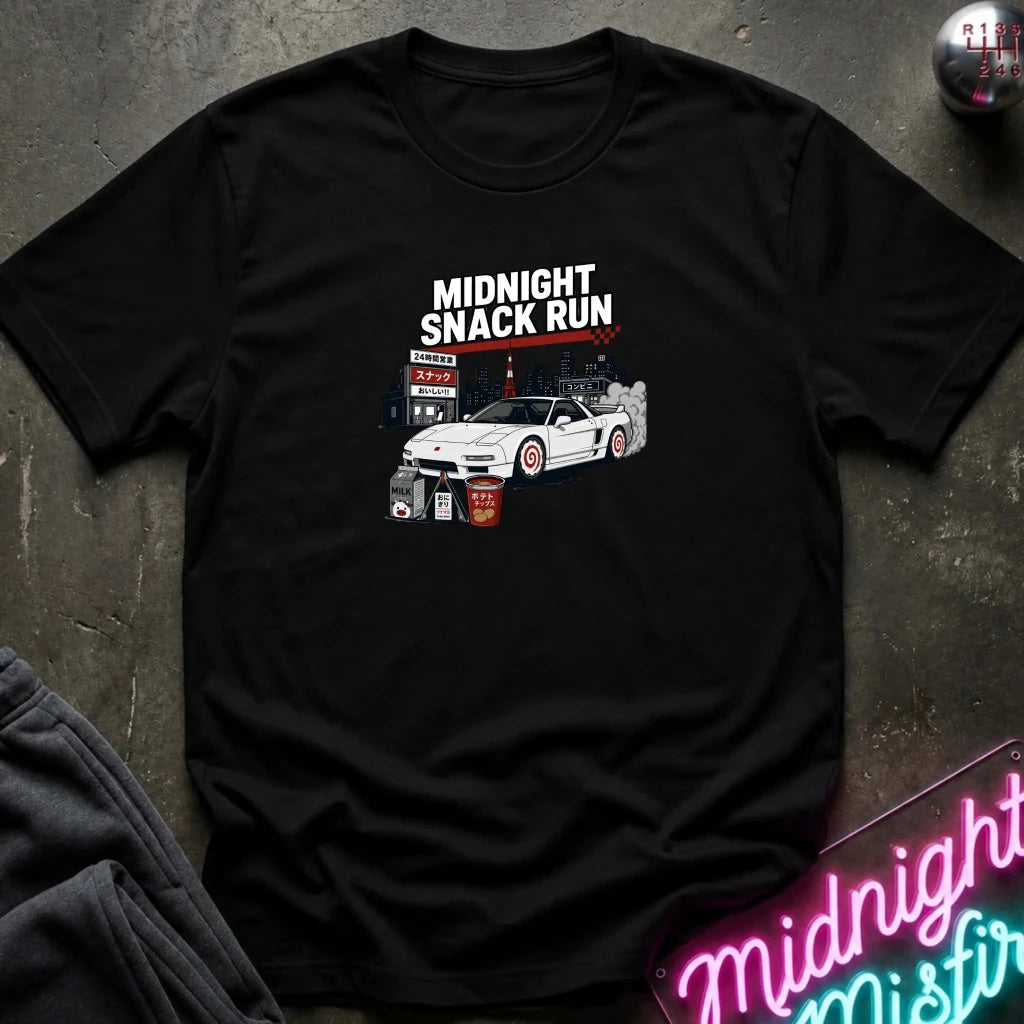 A black short-sleeve graphic t-shirt laid flat on a dark textured surface. The graphic features the text 'MIDNIGHT SNACK RUN' above an illustration of a white sports car with red wheels, a gas station sign, and a smoke effect.
