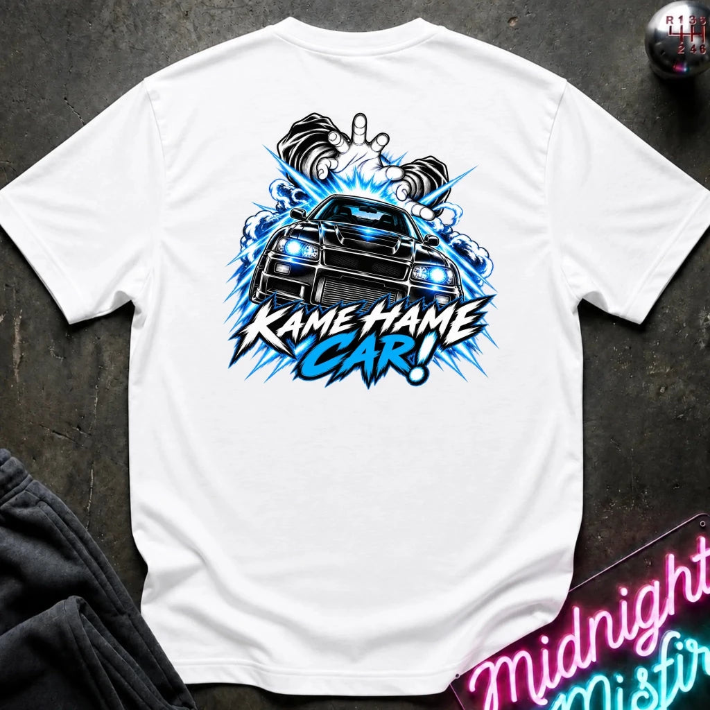 A white short-sleeve t-shirt laid flat on a dark textured surface, featuring a large graphic print of a black sports car with glowing blue headlights and the text 'KAME HAME CAR!' in blue and black. 
