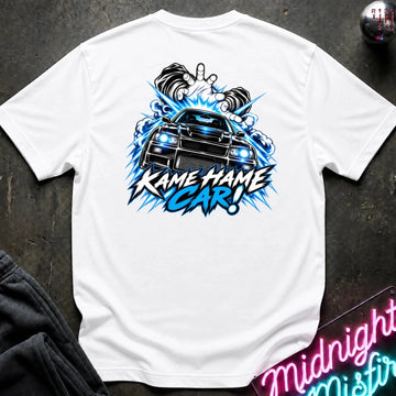 A white short-sleeve t-shirt laid flat on a dark textured surface, featuring a large graphic print of a black sports car with glowing blue headlights and the text 'KAME HAME CAR!' in blue and black. 