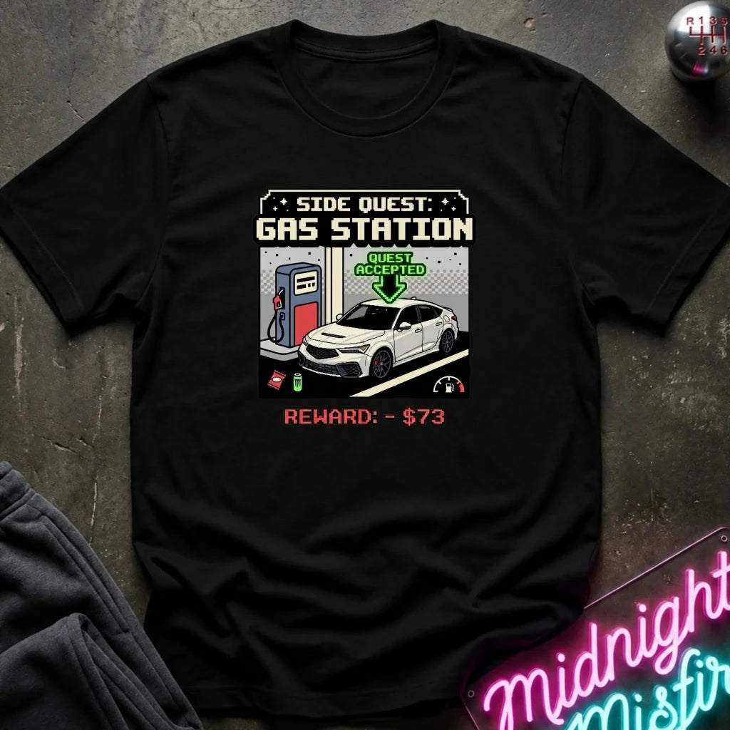 A black short-sleeve graphic t-shirt laid flat on a dark textured surface. The shirt features a retro video game style chest print reading 'SIDE QUEST: GAS STATION' with an illustration of a white car at a gas pump and 'REWARD: - $73' in red text.