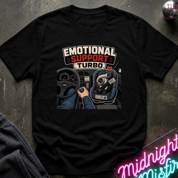 A black short-sleeve graphic t-shirt laid flat on a dark textured surface. The shirt features a graphic print with the words 'EMOTIONAL SUPPORT TURBO' above an illustration of a hand on a steering wheel inside a car.