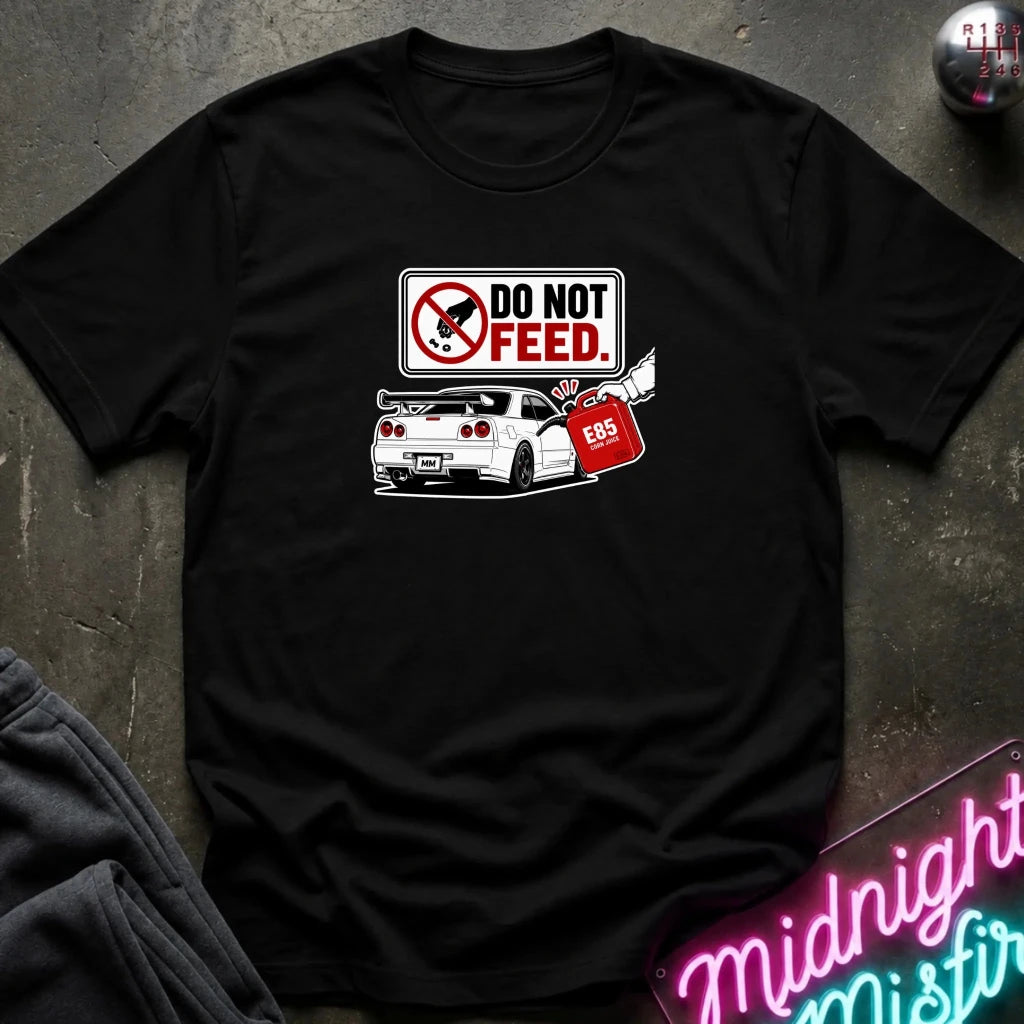 A black short-sleeve graphic t-shirt laid flat on a dark textured surface. The graphic features a 'DO NOT FEED' sign.