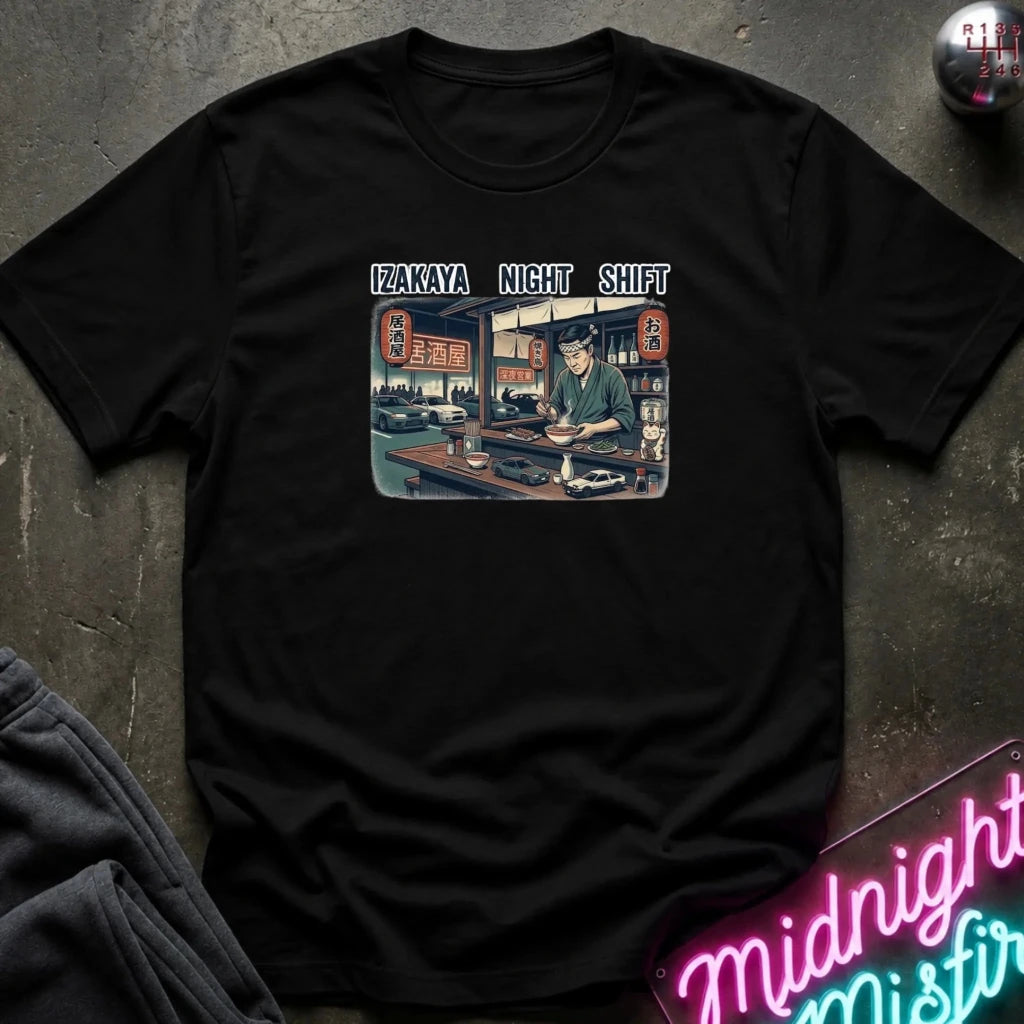 A black short-sleeve graphic t-shirt laid flat on a dark textured surface. The graphic features the text 'IZAKAYA NIGHT SHIFT' above a retro-style illustration of a worker at a counter.