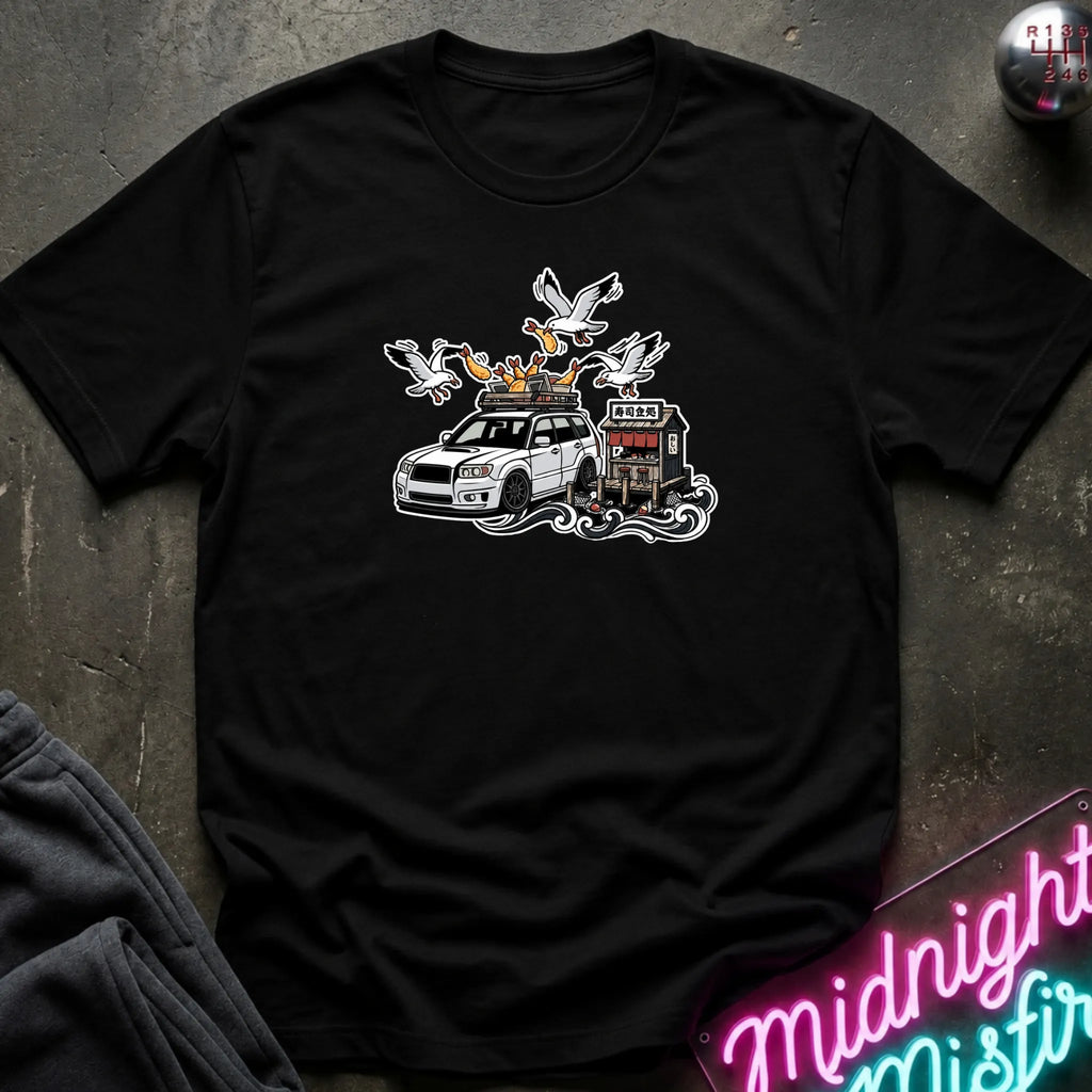 A black short-sleeve graphic t-shirt laid flat on a dark textured surface. The graphic features a white station wagon with a roof rack carrying seagulls and a small building.