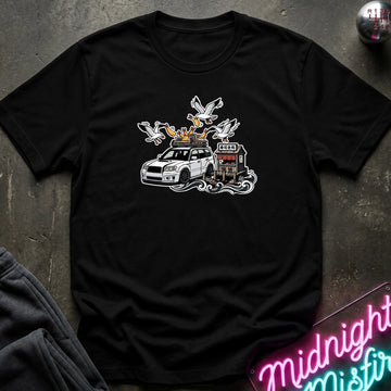 A black short-sleeve graphic t-shirt laid flat on a dark textured surface. The graphic features a white station wagon with a roof rack carrying seagulls and a small building.