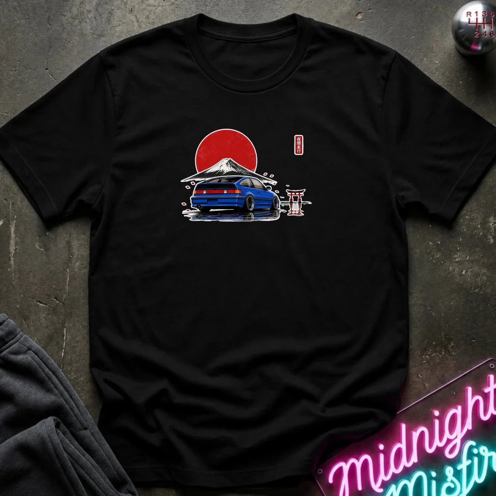A black short-sleeve t-shirt laid flat on a dark textured surface, featuring a graphic print of a blue sports car in front of a red sun and mountain, with a small red vertical text element. A neon sign and folded dark fabric are partially visible around the edges.