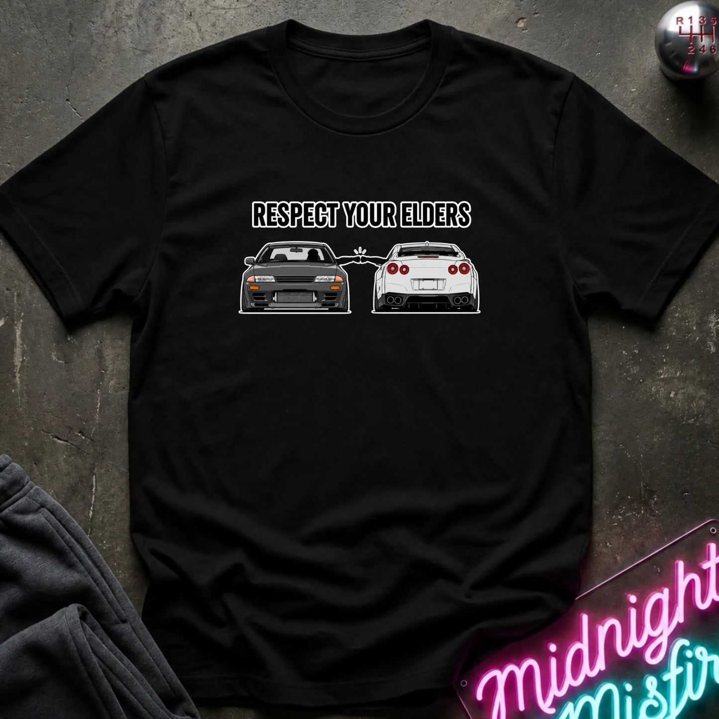 A black short-sleeve graphic T-shirt laid flat on a dark textured surface. The shirt has a black printed slogan reading 'RESPECT YOUR ELDERS' above two cars, with a neon sign and dark clothing partially visible around it.