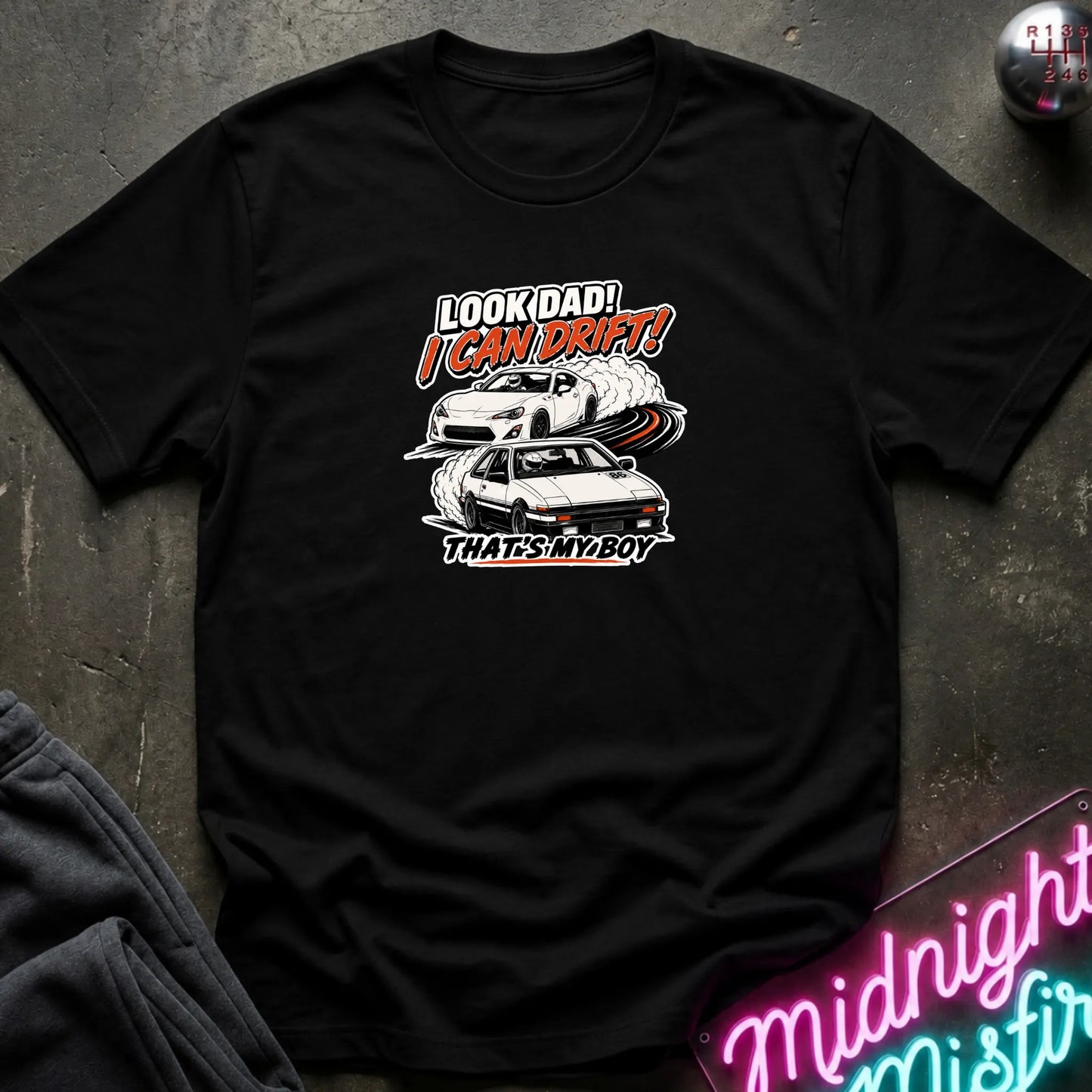 A black  short-sleeved t-shirt  that has a cars drifting which are older and newer generations of toyotas.