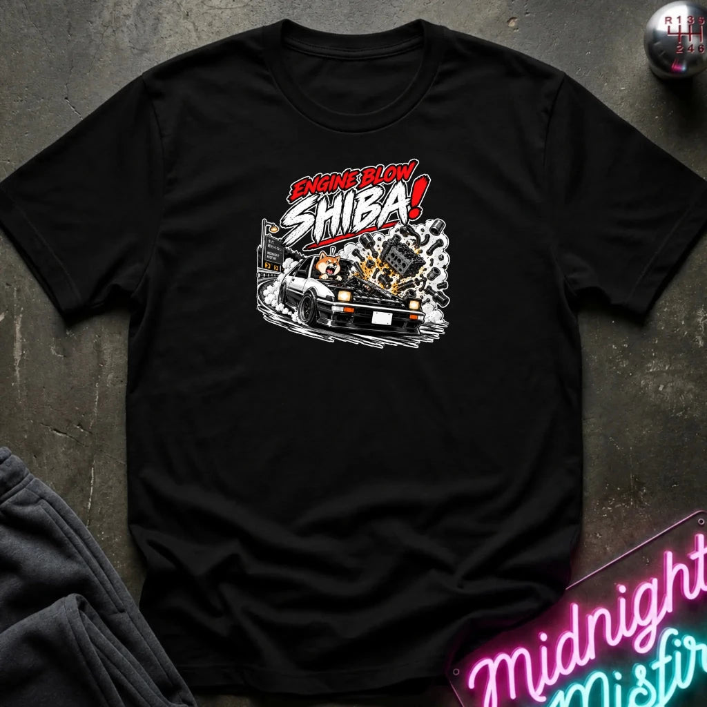 A black short-sleeve graphic t-shirt laid flat on a dark textured surface. The shirt features a stylized graphic of a white sports car with a glowing engine and the text 'ENGINE BLOW SHIBA!' in red, white, and black.
