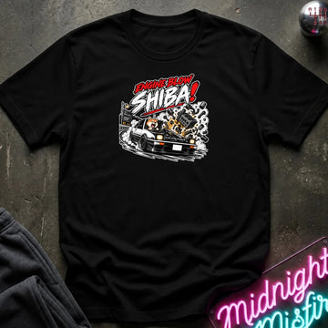 A black short-sleeve graphic t-shirt laid flat on a dark textured surface. The shirt features a stylized graphic of a white sports car with a glowing engine and the text 'ENGINE BLOW SHIBA!' in red, white, and black.