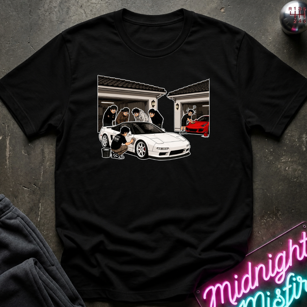 A black short-sleeve crewneck t-shirt laid flat on a dark textured surface. The shirt features a graphic print of a white sports car in front of a house with people and a red car in the garage.