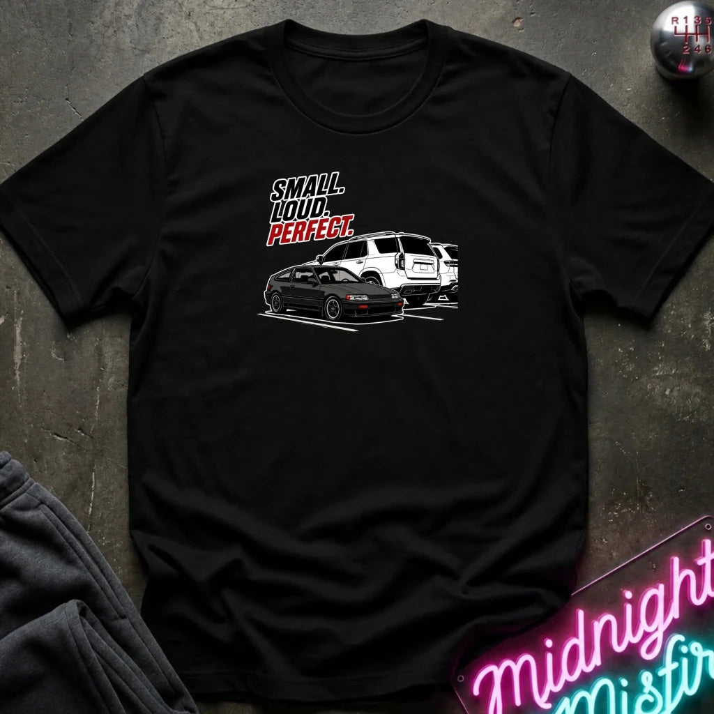 A black short-sleeve graphic t-shirt laid flat on a dark textured surface. The graphic features the text 'SMALL louD PERFECT.