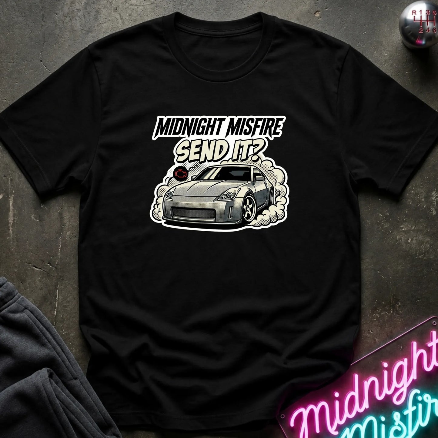 A black short-sleeve graphic t-shirt laid flat on a dark textured surface. The shirt features a graphic of a silver sports car with smoke and the text 'MIDNIGHT MISFIRE SEND IT?' in white and black.