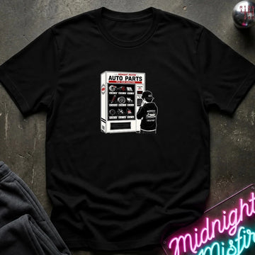 A black short-sleeve t-shirt with a graphic print of an 'Auto Parts' vending machine and a person, laid flat on a dark textured surface with a neon sign and sweatpants partially visible.
