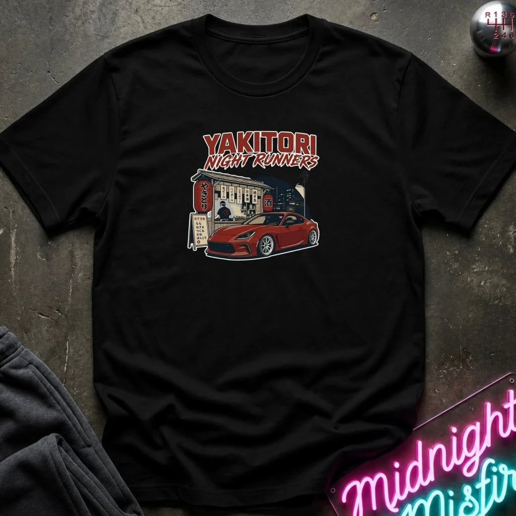 A black short-sleeve graphic t-shirt laid flat on a dark textured surface. The graphic features the text 'YAKITORI NIGHT RUNERS' in red and white above an illustration of a red sports car parked outside a building.