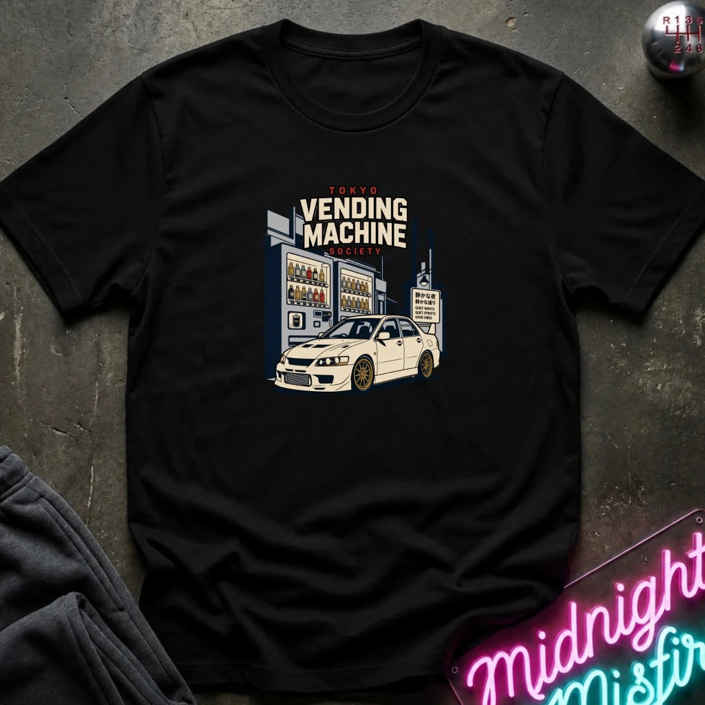 A black short-sleeve graphic T-shirt laid flat on a dark textured surface. The shirt features a central graphic of a white sports car in front of vending machines with the text 'Tokyo Vending Machine Society'.