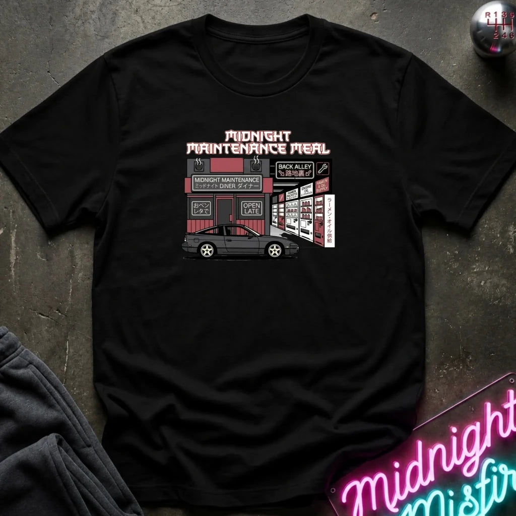 A black short-sleeve graphic t-shirt laid flat on a dark textured surface. The graphic features a retro-style illustration of a garage with a black car in front, and the text 'MIDNIGHT MAINTENANCE MEAL' in pink and white.