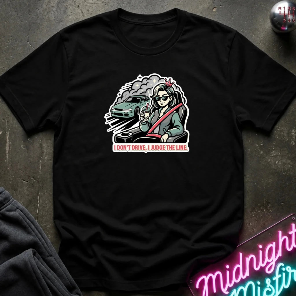 A black short-sleeve graphic t-shirt laid flat on a dark textured surface. The graphic shows a woman in a green car with the text 'I DON'T DRIVE, I JUDGE THE LINE.