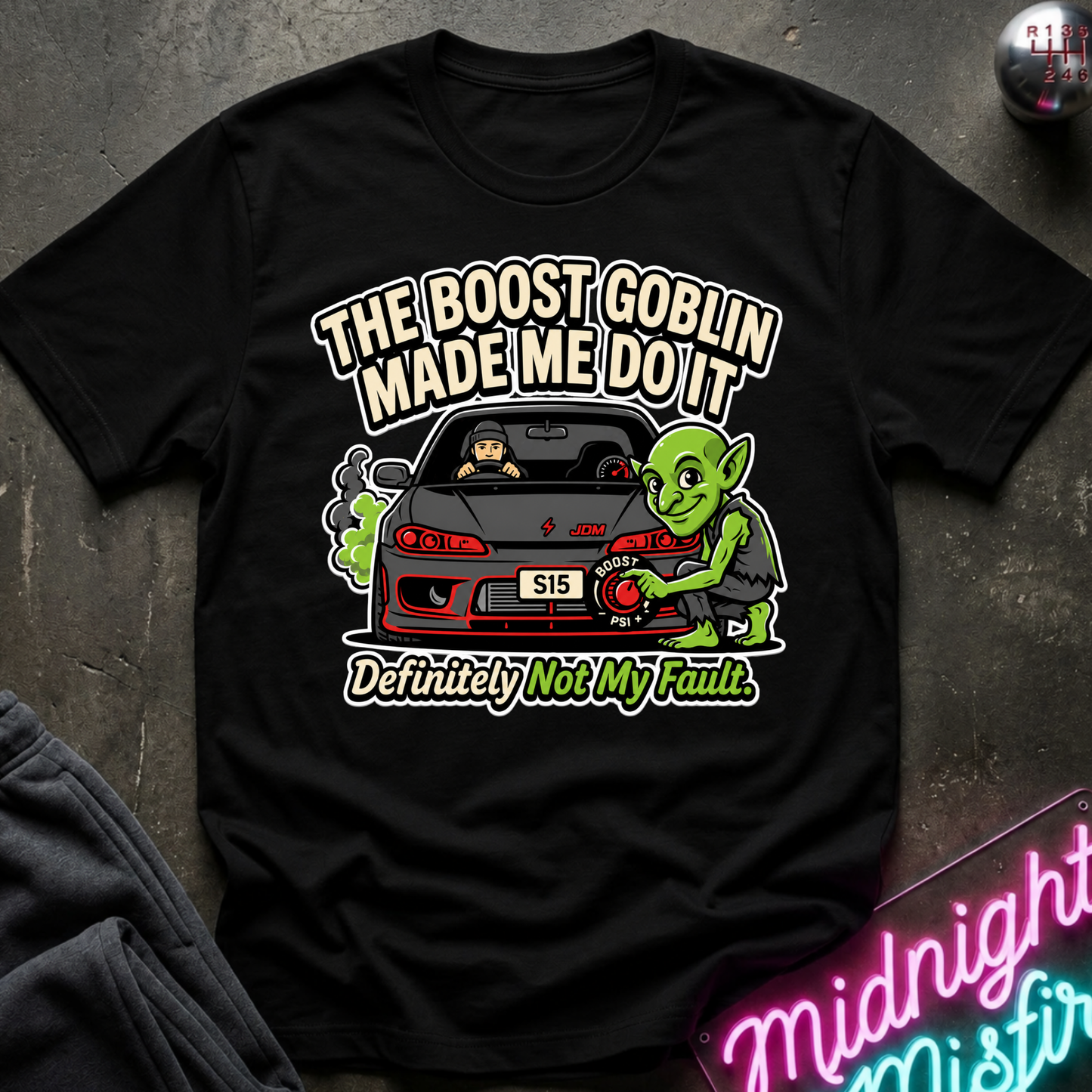A black short-sleeve graphic T-shirt laid flat on a dark textured surface. The shirt features a colorful cartoon print of a green Yoda-like character beside a red car, with the text 'THE BOOST GOBLIN MADE ME DO IT' and 'Definitely Not My Fault.