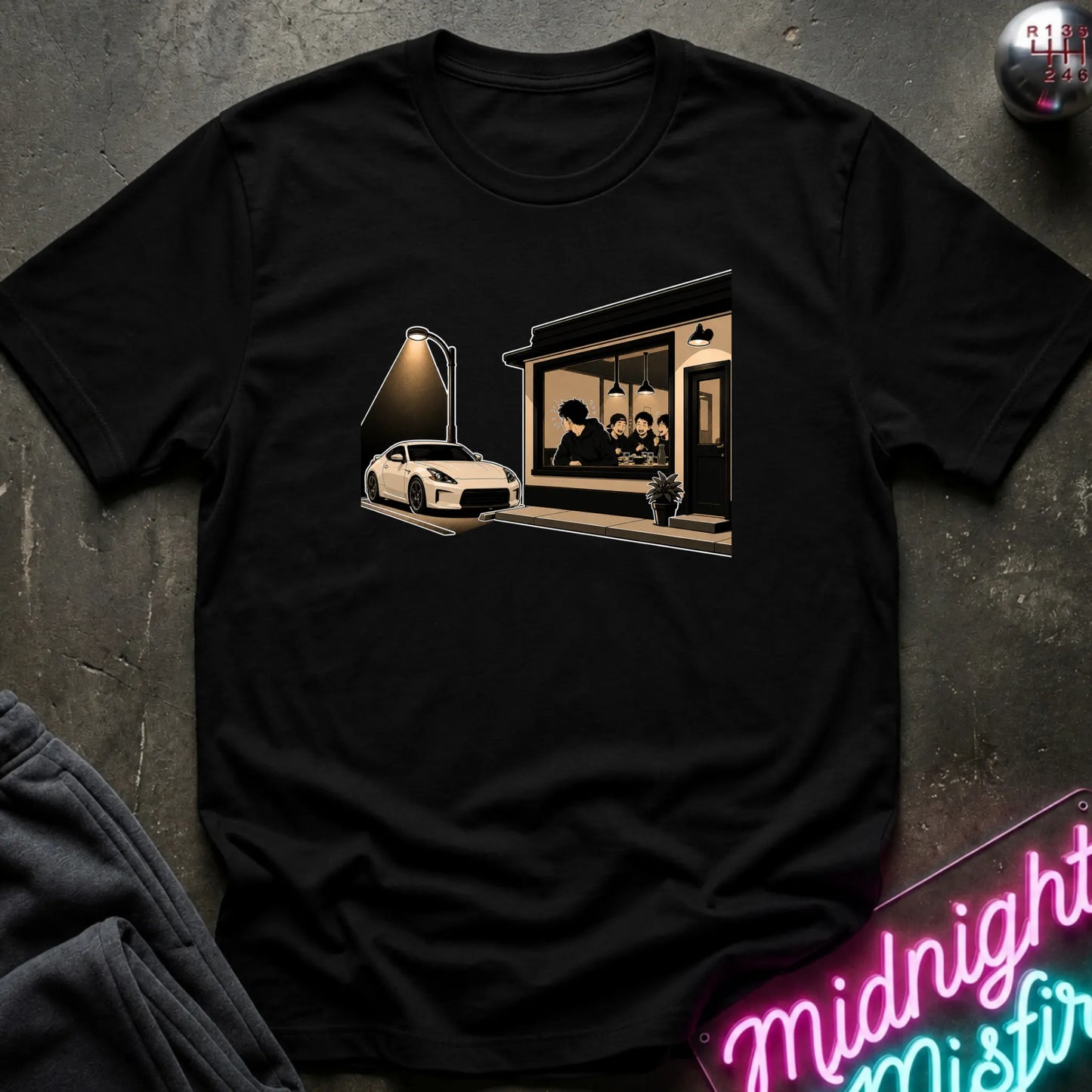 A black short-sleeve graphic t-shirt laid flat on a dark textured surface. The graphic shows a white sports car parked outside a dimly lit cafe with a neon sign in the foreground.