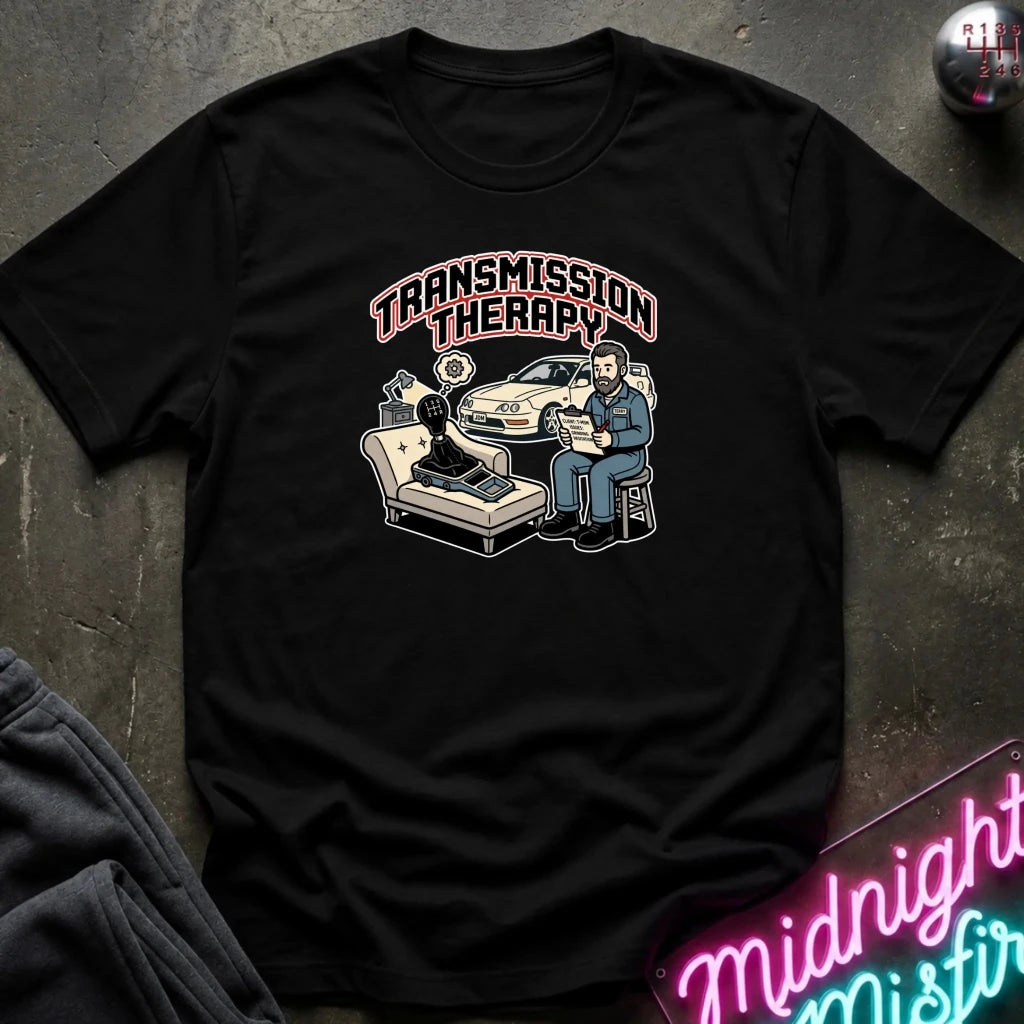 A black short-sleeve graphic t-shirt laid flat on a dark textured surface. The shirt features a retro-style illustration of a therapist sitting next to a patient on a chaise lounge with a car in the background, under the text 'TRANSMISSION THERAPY' in pink and white.