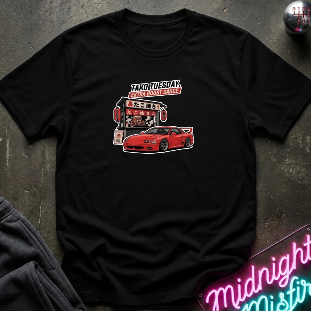 A black short-sleeve graphic t-shirt laid flat on a dark textured surface. The graphic features a red sports car and a food stall with Japanese text reading 'TAKOTUESDAY' 