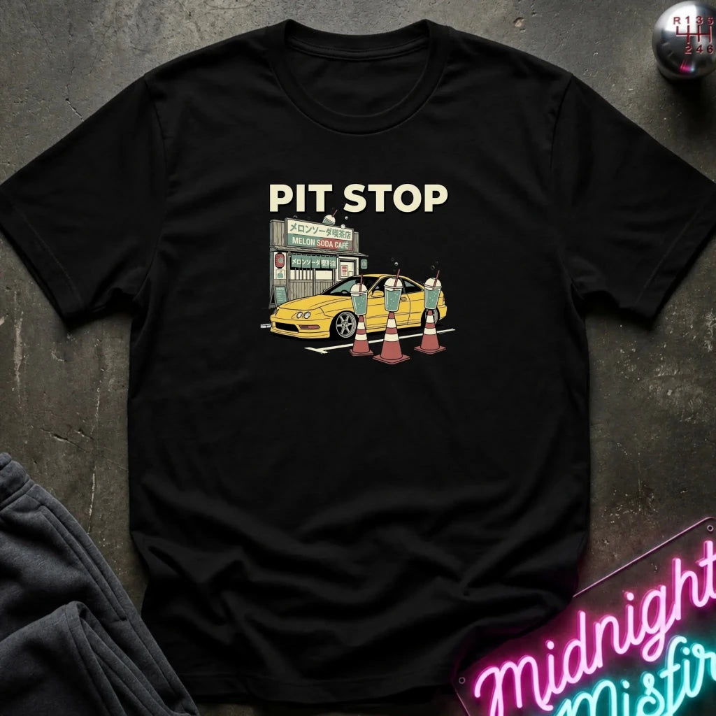 A black short-sleeve graphic t-shirt laid flat on a dark textured surface. The graphic features the words 'PIT STOP' in cream above an illustration of a yellow sports car at a Japanese-style gas station with traffic cones and two figures.