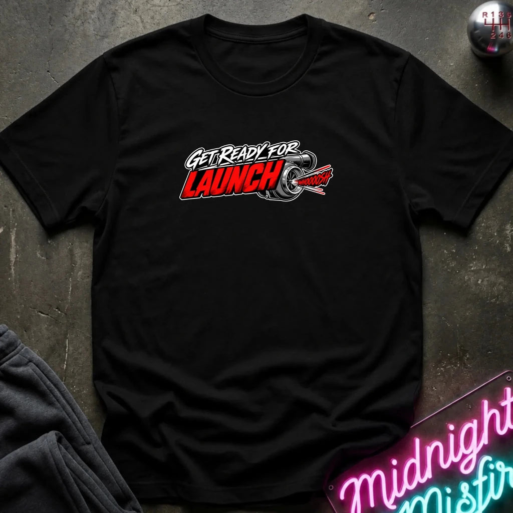 A black short-sleeve crewneck t-shirt with a 'GET READY FOR LAUNCH' graphic on the chest, styled flat on a dark textured surface with a neon sign and dark pants in the background.