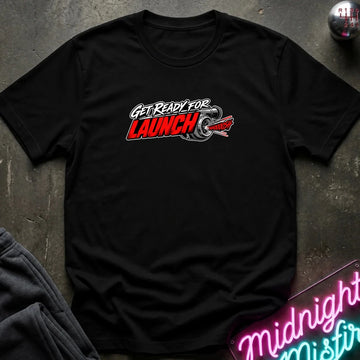 A black short-sleeve crewneck t-shirt with a 'GET READY FOR LAUNCH' graphic on the chest, styled flat on a dark textured surface with a neon sign and dark pants in the background.