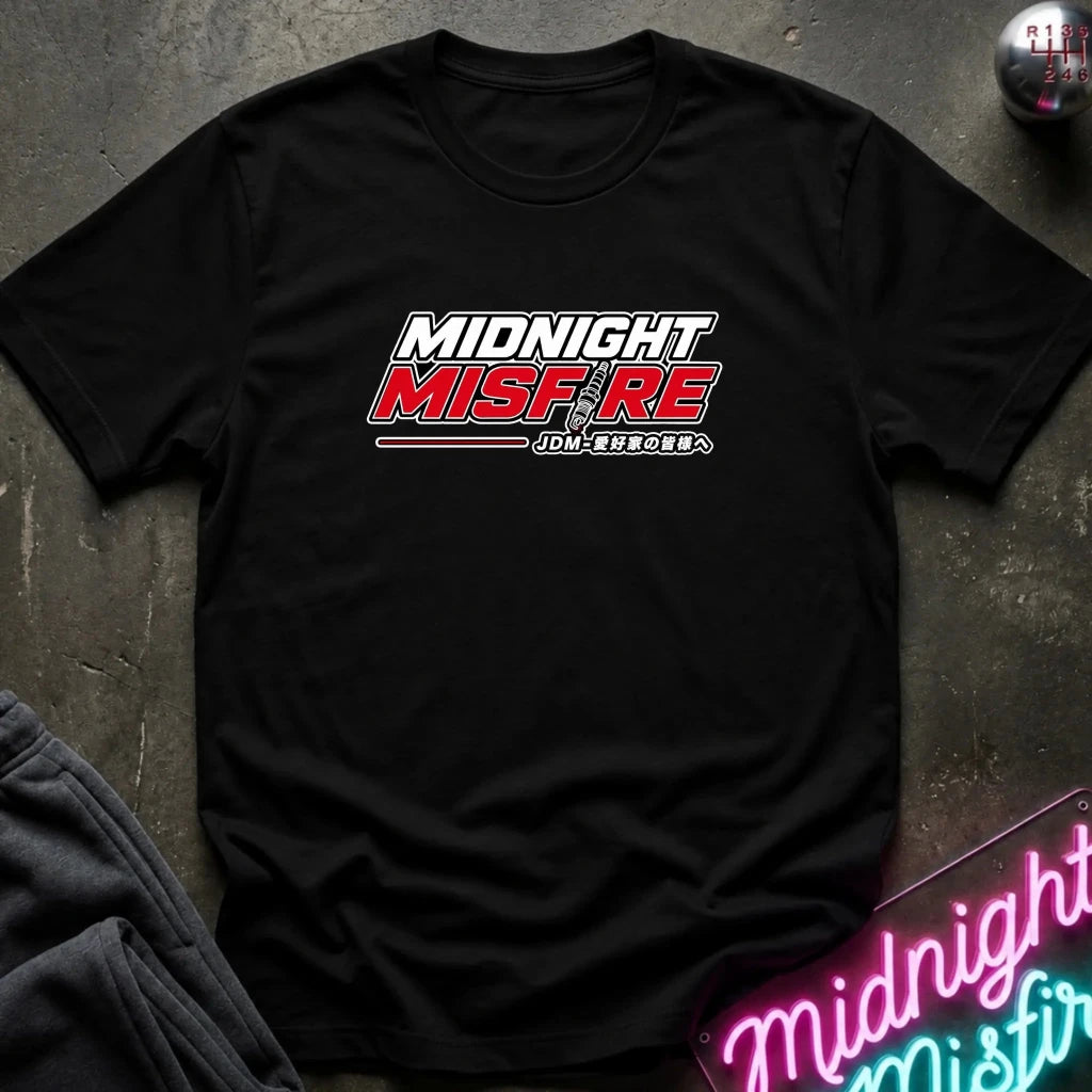 A black short-sleeve t-shirt laid flat on a dark textured surface, featuring a 'MIDNIGHT MISFIRE' graphic on the chest with stylized white and red lettering. A neon sign and folded dark clothing are partially visible in the background as props.