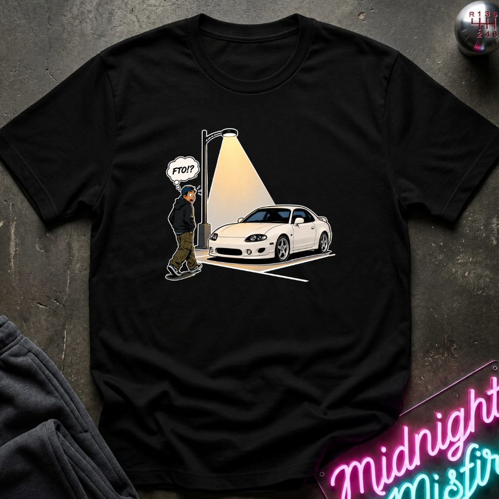 A black short-sleeve graphic t-shirt laid flat on a dark textured surface. The graphic shows a white sports car parked under a streetlamp with a figure walking toward it.