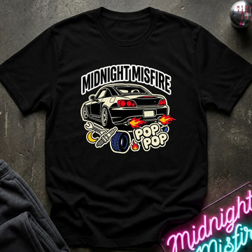A black short-sleeve graphic t-shirt laid flat on a dark textured surface. The shirt features a graphic of a black sports car with flames, a tire, and the text 'MIDNIGHT MISFIRE' and 'POP POP'.