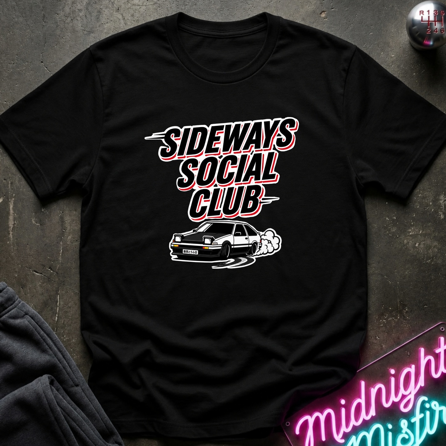A black short-sleeve graphic t-shirt laid flat on a dark textured surface. The shirt features a white and red graphic print that reads 'SIDEWAYS SOCIAL CLUB' above a white car illustration.